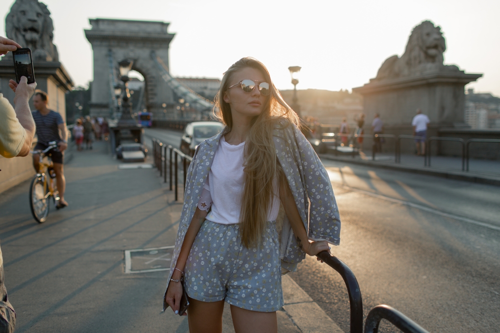 a woman stands by the road in shorts, a t shirt, and blazer. she's wearing sunglasses and there is a bridge in the background, what to wear in budapest