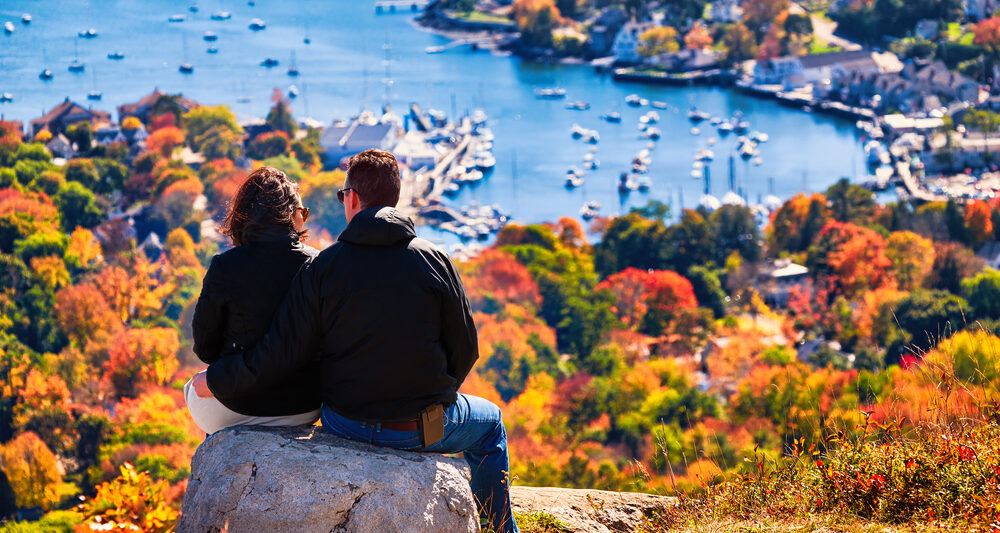 man and woman sitting on top of a mountain with fall foliage in the foreground. they are looking down at the town of Bar Harbor, Maine, with the ocean in the background and some small boats in the water
