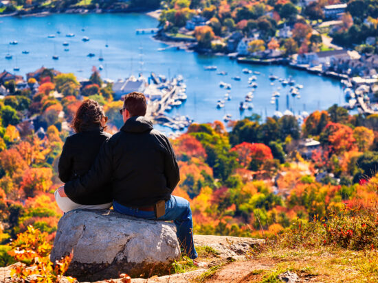man and woman sitting on top of a mountain with fall foliage in the foreground. they are looking down at the town of Bar Harbor, Maine, with the ocean in the background and some small boats in the water