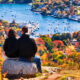 man and woman sitting on top of a mountain with fall foliage in the foreground. they are looking down at the town of Bar Harbor, Maine, with the ocean in the background and some small boats in the water