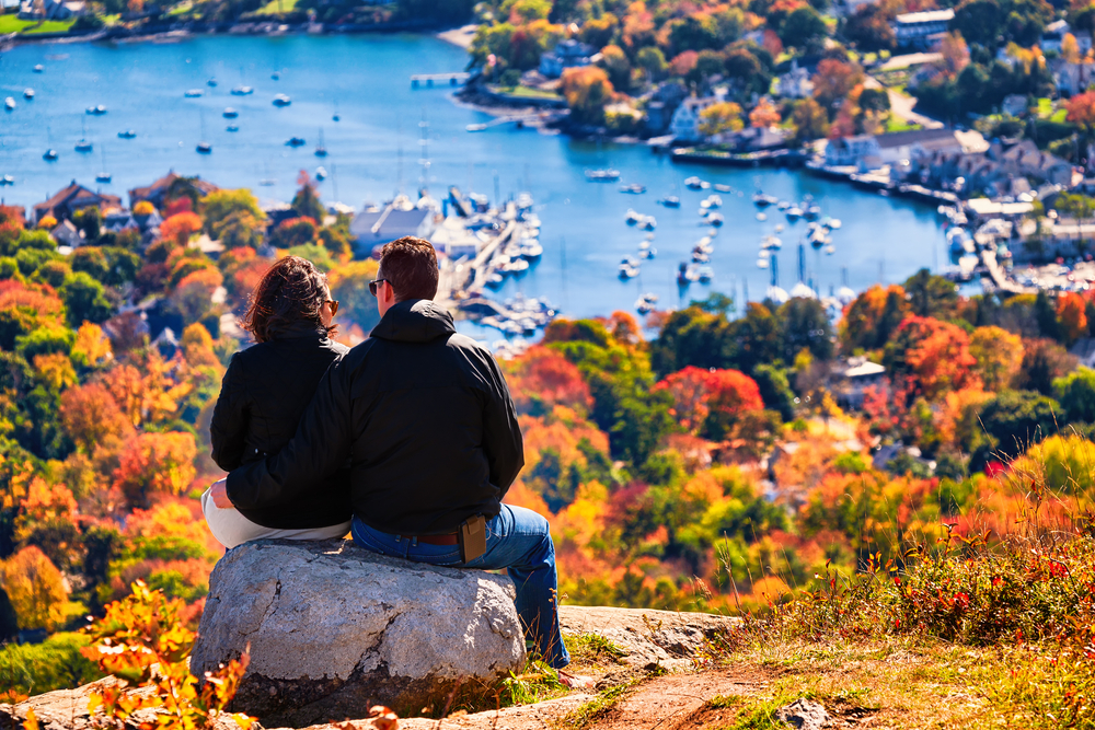 man and woman sitting on top of a mountain with fall foliage in the foreground. they are looking down at the town of Bar Harbor, Maine, with the ocean in the background and some small boats in the water