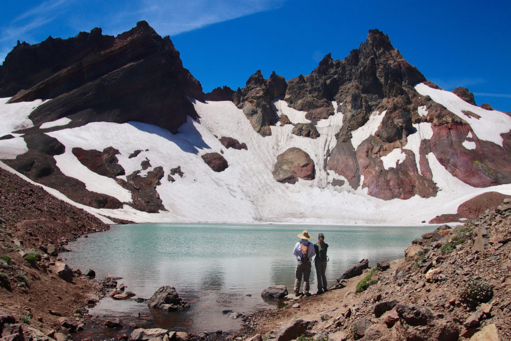 No Name Lake, Bend Glacier & a couple stands at base of lake with snow covered mountains in backdrop