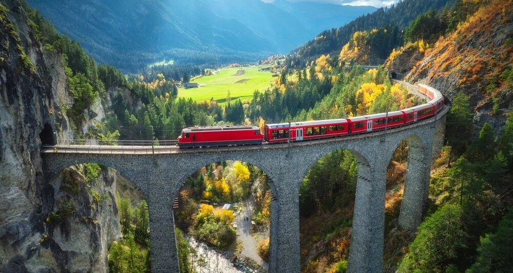 a scenic train ride in europe crossing a viaduct. the train is red and it is going across a very tall bridge meant for trains. there are green mountains and fields around it, and the sky has clouds and a little blue