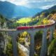 a scenic train ride in europe crossing a viaduct. the train is red and it is going across a very tall bridge meant for trains. there are green mountains and fields around it, and the sky has clouds and a little blue