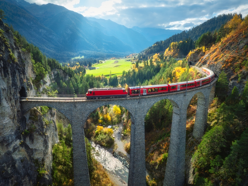 Aerial view of modern red train on Landwasser viaduct in alpine mountains on the Bernina Express. the train is red and it is going across a very tall bridge meant for trains. there are green mountains and fields around it, and the sky has clouds and a little blue