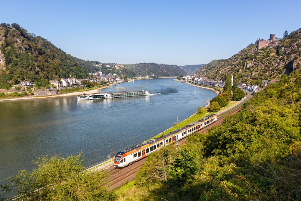 THe Rhine Valley train passing along the Rhine River where you can see river boats and castles int he background