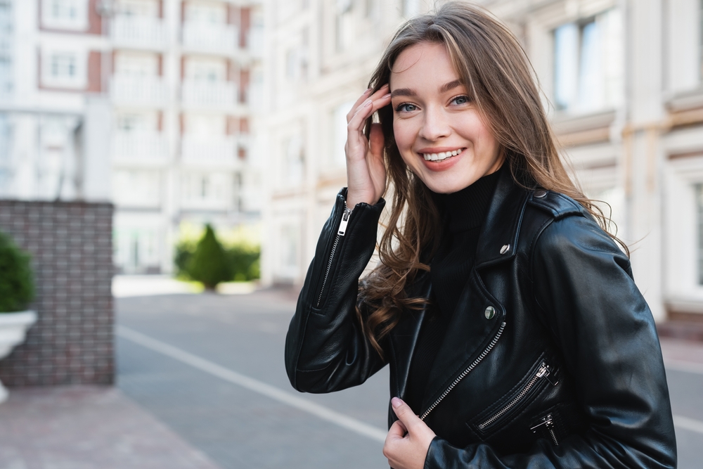 A woman brushes her hair out of her face and smiles at the camera while wearing a black turtleneck and leather jacket.