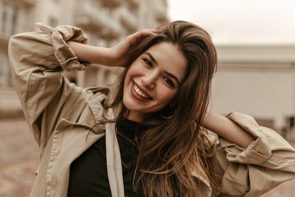 A woman smiles at the camera, knowing what to wear in Italy in November with her hoop earrings trench coat jacket and thick blacks shirt.