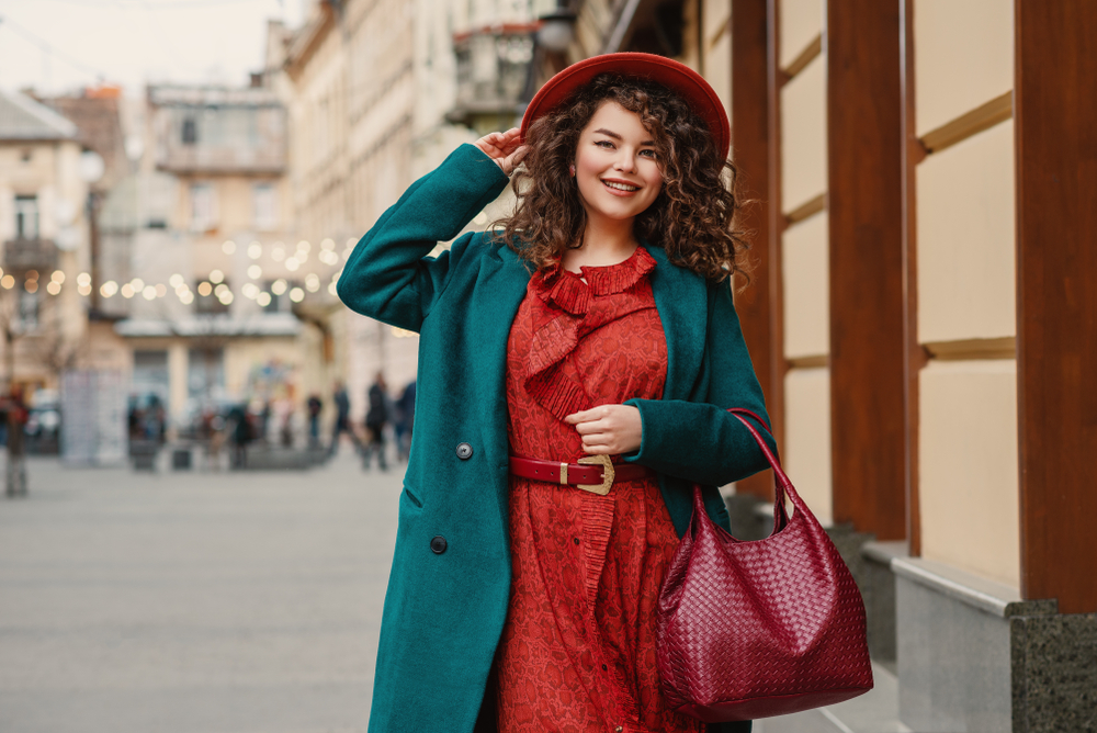 A woman with curly hair adjusts her hat that matches her red dress as she walks through, wearing her blue statement coat.