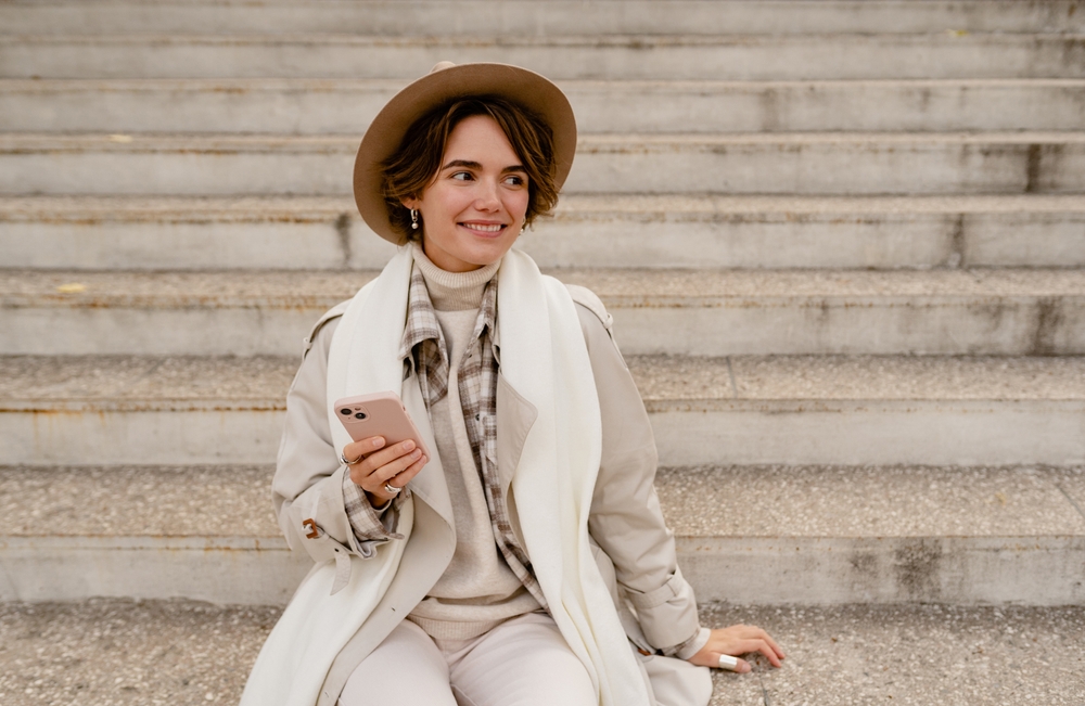 A woman with a short haircut and a hat sits on the stairs of a building, holding her phone, wearing a waterproof jacket, scarf and more.