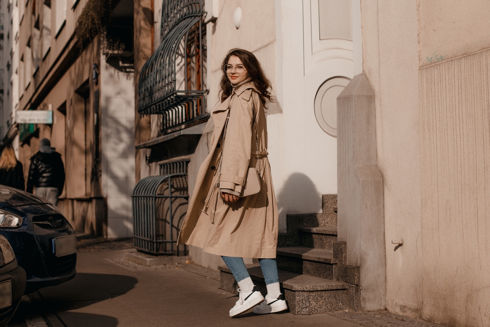 A woman knows what to wear in Italy in November from a casual point of view in her long trench, crossbody, slim jeans and trainers.