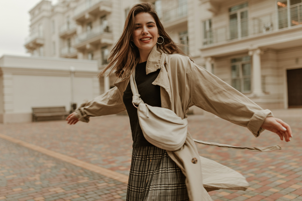 A woman twirls with her arms out in a cobblestone street, knowing what to wear in Italy in November, in her plaid skirt, crossbody bag and trench.