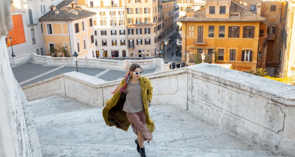 A woman knows what to wear in Italy in November as she climbs some stairs in her boots, skirt, thick sweater and layered trench coat.