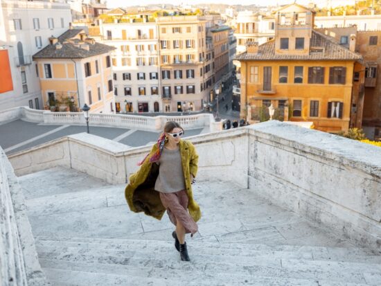 A woman knows what to wear in Italy in November as she climbs some stairs in her boots, skirt, thick sweater and layered trench coat.