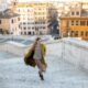 A woman knows what to wear in Italy in November as she climbs some stairs in her boots, skirt, thick sweater and layered trench coat.