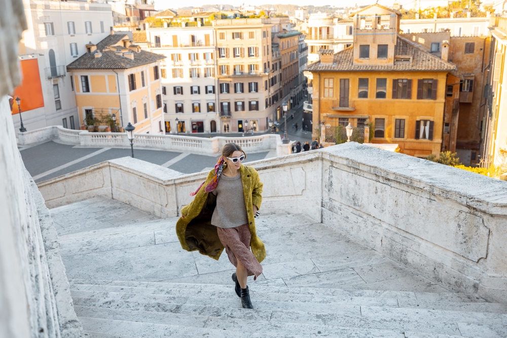 A woman knows what to wear in Italy in November as she climbs stairs in a wool coat, sweater, skirt and boots.