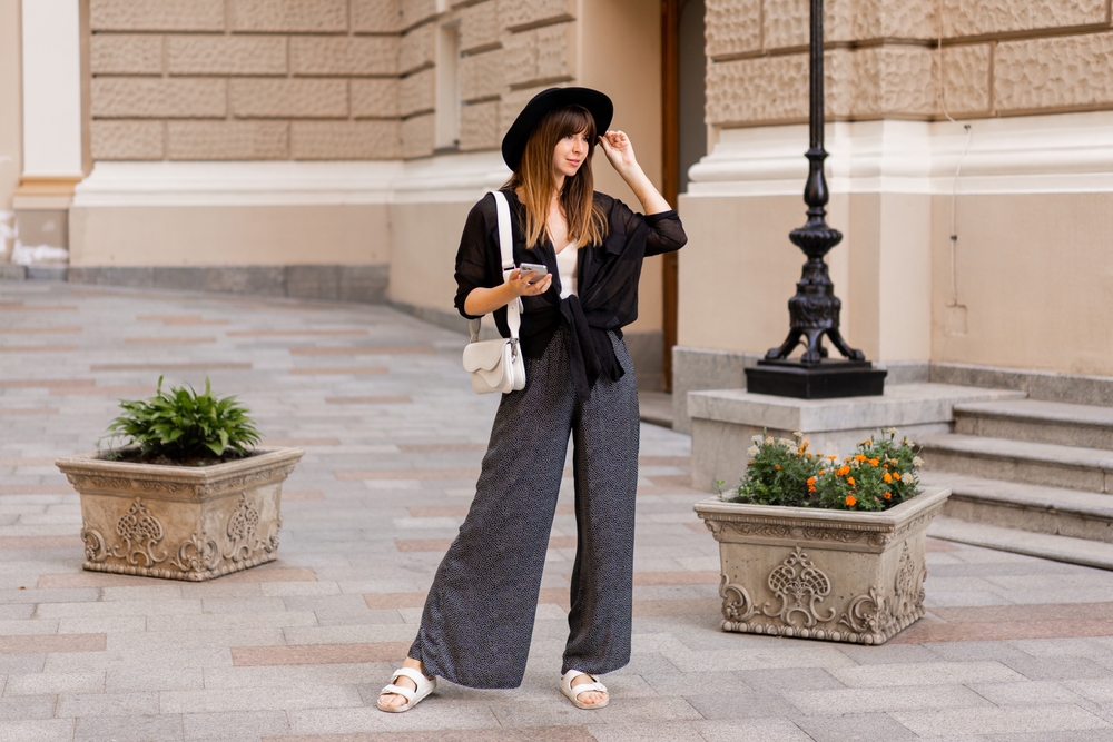 The woman in this photo poses on the street knowing what to wear in Italy in November in her flowy pants, sheer top, and hat.