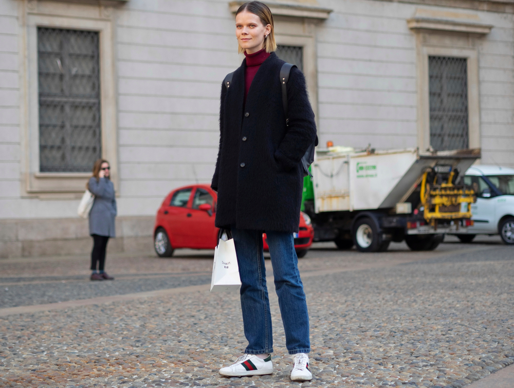 A woman smiles while standing on cobblestone streets, wearing a turtleneck, cardigan, jeans and trainers.