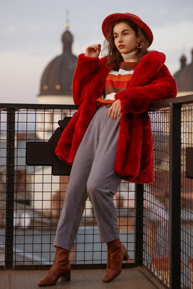 A young girl leans against a corner fence, serving looks as she knows what to wear in Italy in November for a statement with her houndstooth pants, striped turtleneck and faux fur red coat.