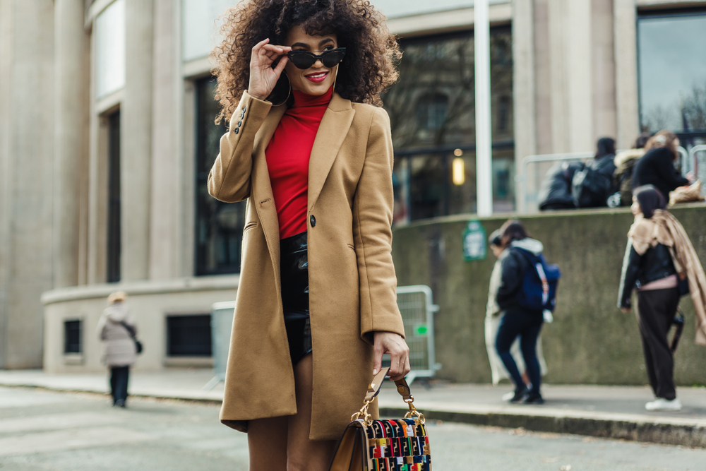 A woman with curly hair looks over her sunglasses as she carries her purse while wearing a skirt, red turtleneck and wool jacket .