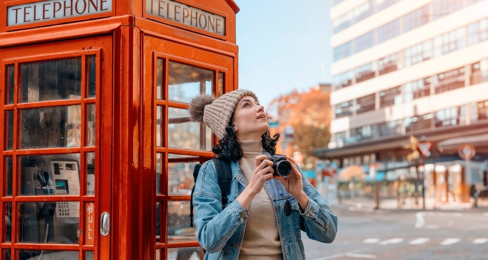 A woman looks up at the sky while leaning by red telephone booth: she knows what to wear in London in November in her turtleneck, matching beanie, jeans and denim jacket.