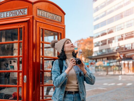 A woman looks up at the sky while leaning by red telephone booth: she knows what to wear in London in November in her turtleneck, matching beanie, jeans and denim jacket.