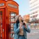 A woman looks up at the sky while leaning by red telephone booth: she knows what to wear in London in November in her turtleneck, matching beanie, jeans and denim jacket.
