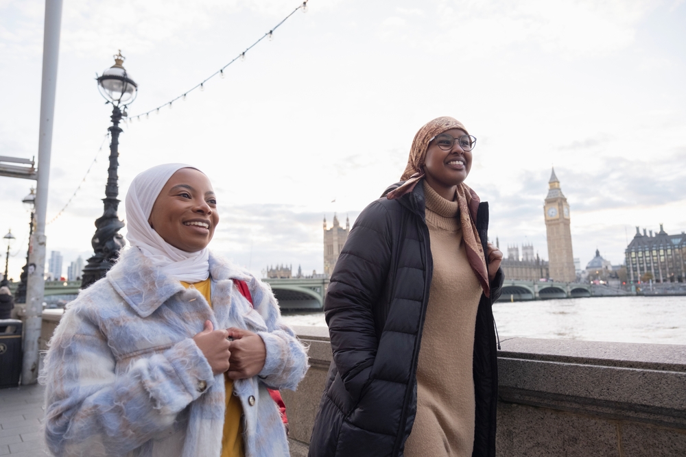 Two friends walk down the River Thames in parkas and thick sweater dresses and shirts, along with head coverings too.