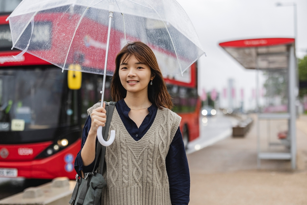 A young Asian girl walks the streets of London in the rain, knowing what to wear in London in November with her black button down, umbrella and knit vest.