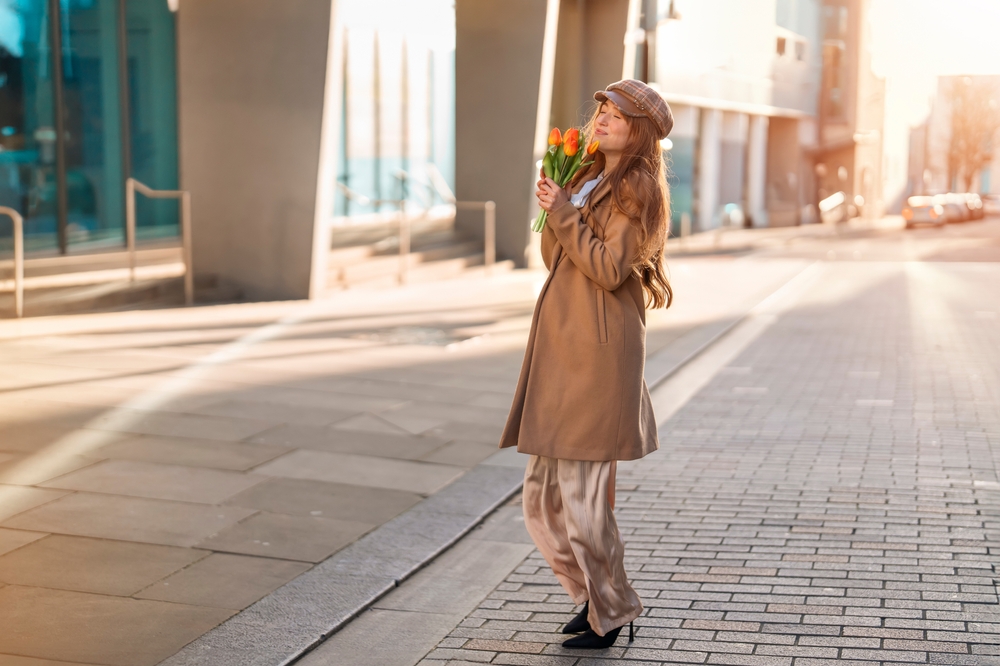 A woman poses on a street in London, knowing what to wear in London in November in her chino pants, long coat, and hat. She smells flowers in her hand!