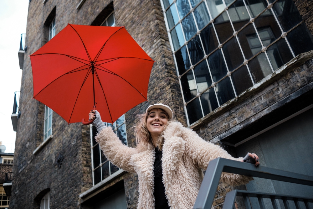 A woman poses on the stairs of a older, stone building in her faux fur coat, gloves, and hat, while also carrying a bright red umbrella.