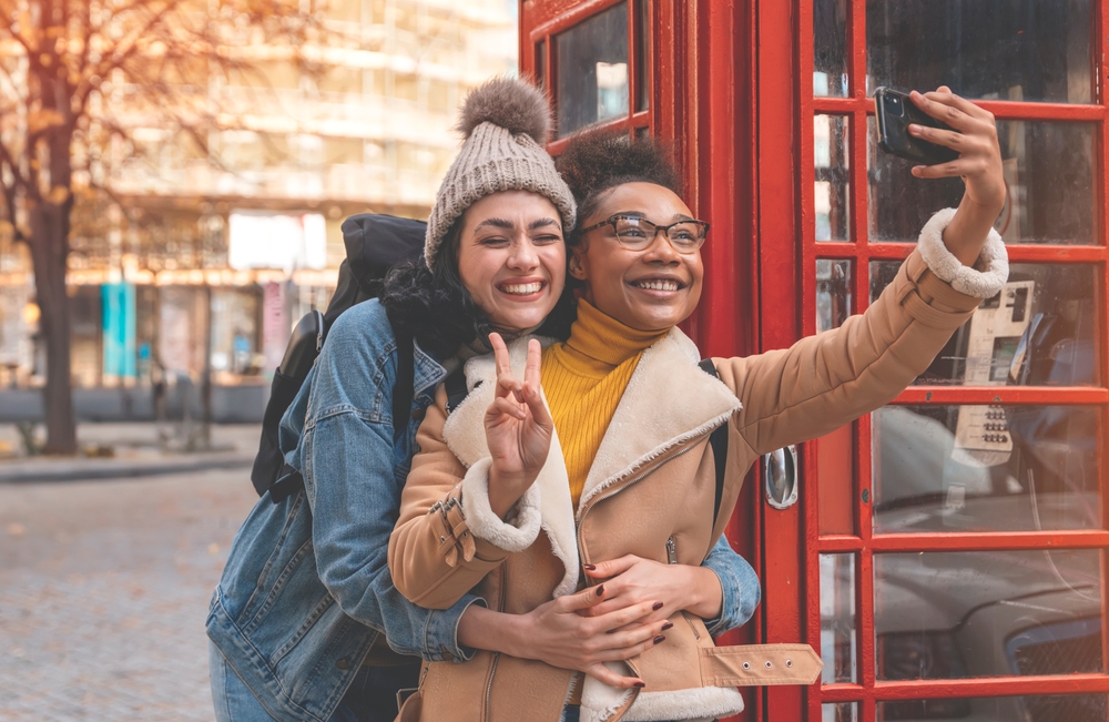 Friends pose for a selfie outside a red photo booth, both in warm jackets and long sleeves to stay warm.