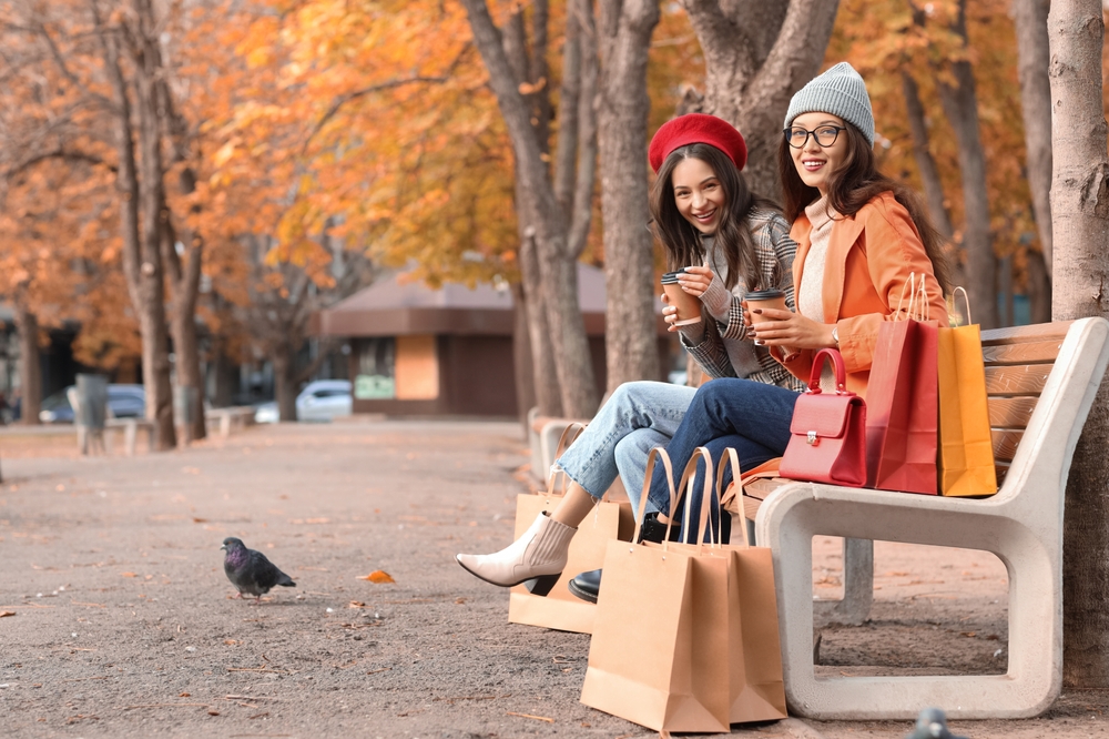 Two friends sit on a bench with shopping bags, both in boots, jeans, beanies, and jackets over turtlenecks: they know what to wear in New York City in November.