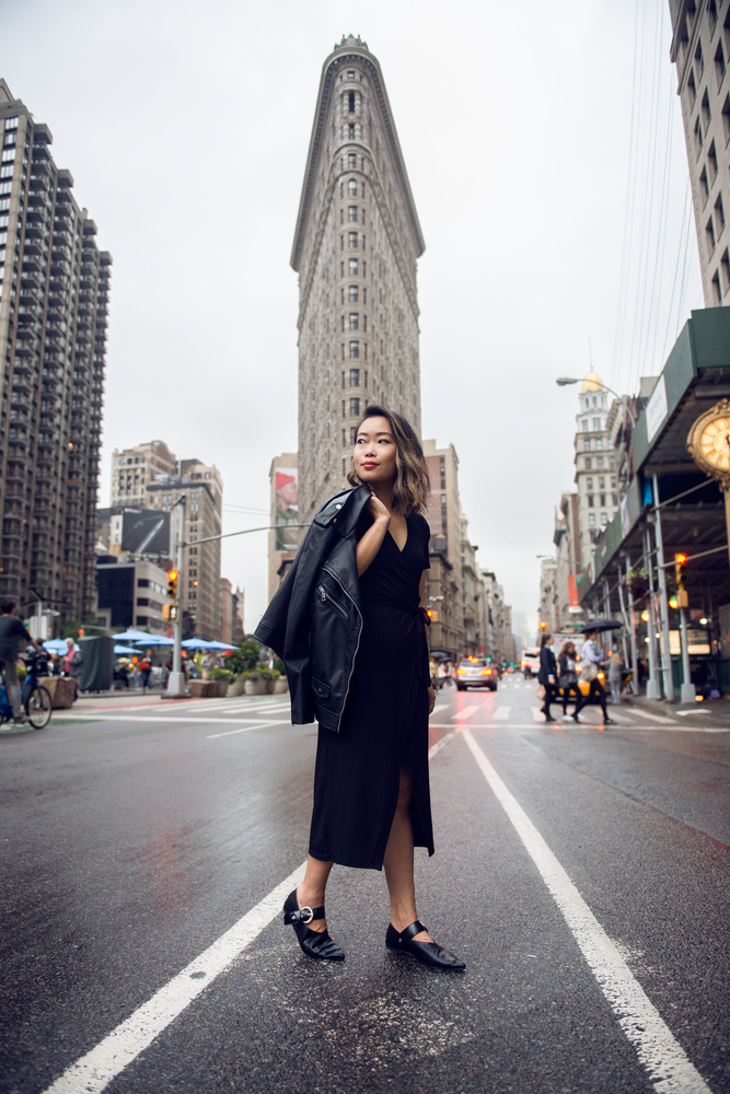 A woman walks through a cool evening in NYC in loafers, a black dress and a leather jacket slung over her shoulder.