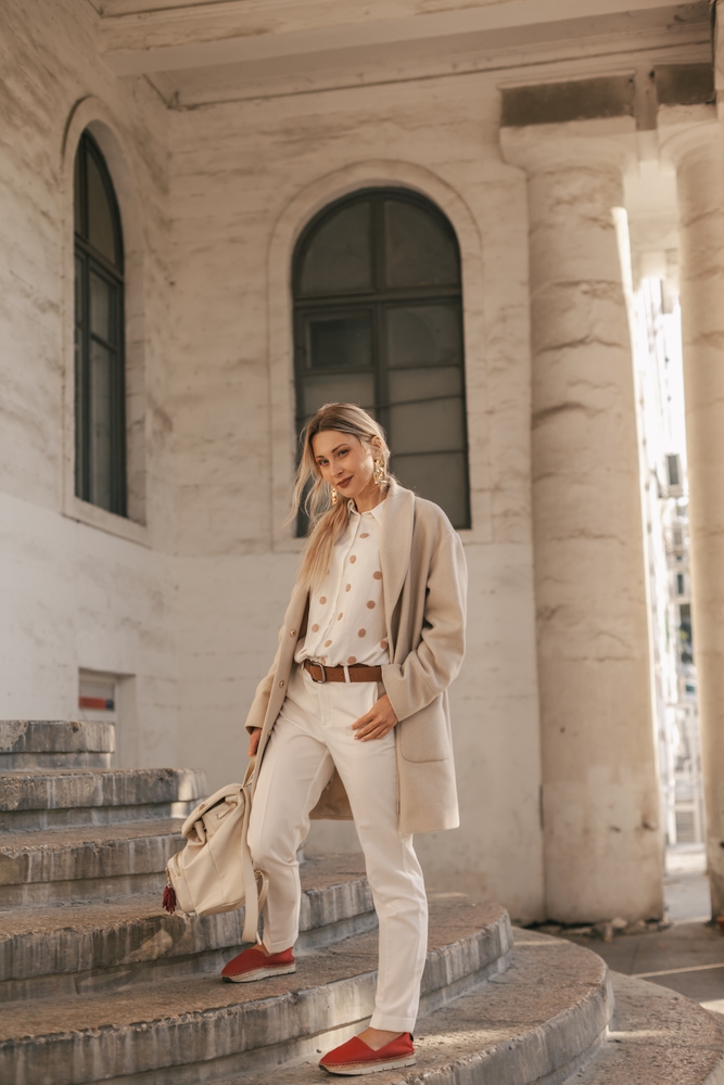 A woman n tailored pants, a silk button down and a wool cardigan poses on stairs while holding her matching bag, knowing full well what to wear in New York City in November.