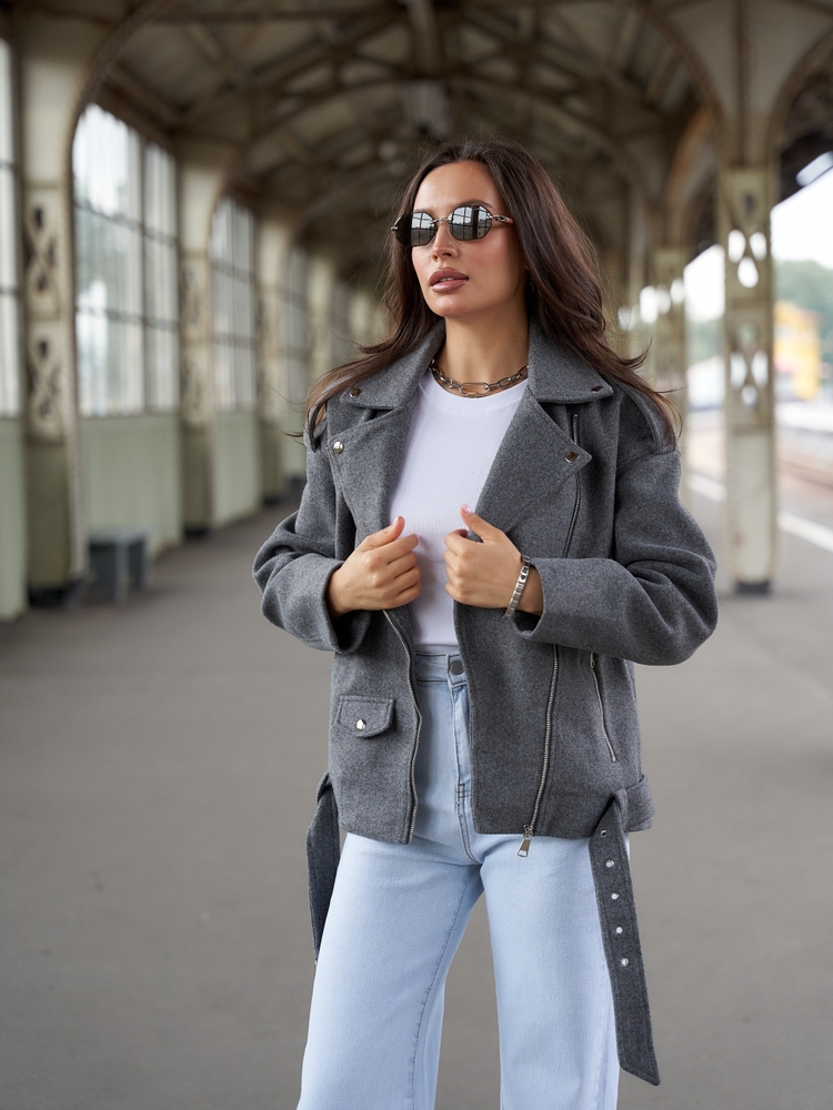 A woman stands and holds her wool jacket over her white t-shirt and jeans: she is comfortable and stylish as she waits for public transport.