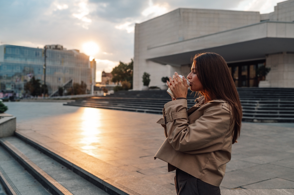 A woman sips her coffee in NYC as the sun rises, her leather jacket over her tailored pants.