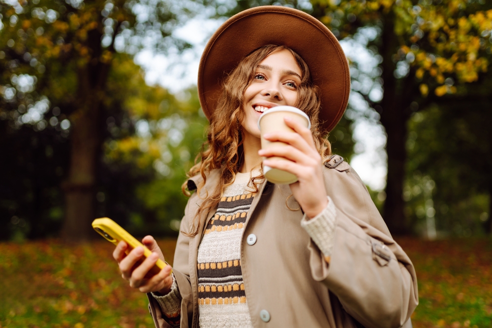 A woman wears a warm hat, a tan trench coat, and a knitted sweater as she holds her phone and drinks a warm coffee.