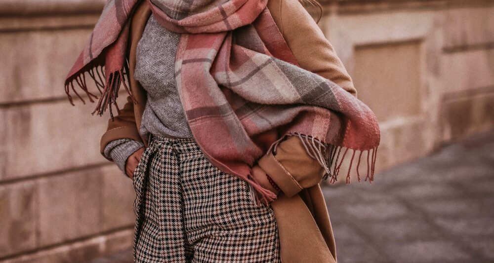 A close up shot of a woman who knows what to wear in New York City in November shows her plaid pants, thick turtle neck, beige jacket and plaid scarf: all fall colors and on theme too!