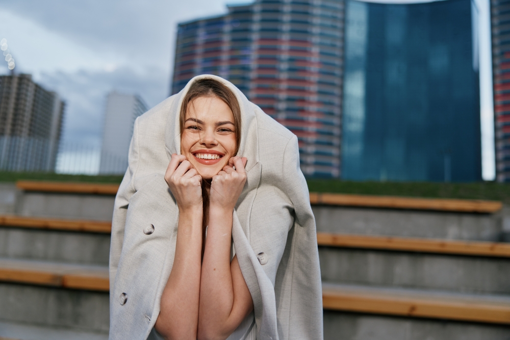 A woman jokingly pulls her trench coat over her head to stay warm, smiling at the camera.
