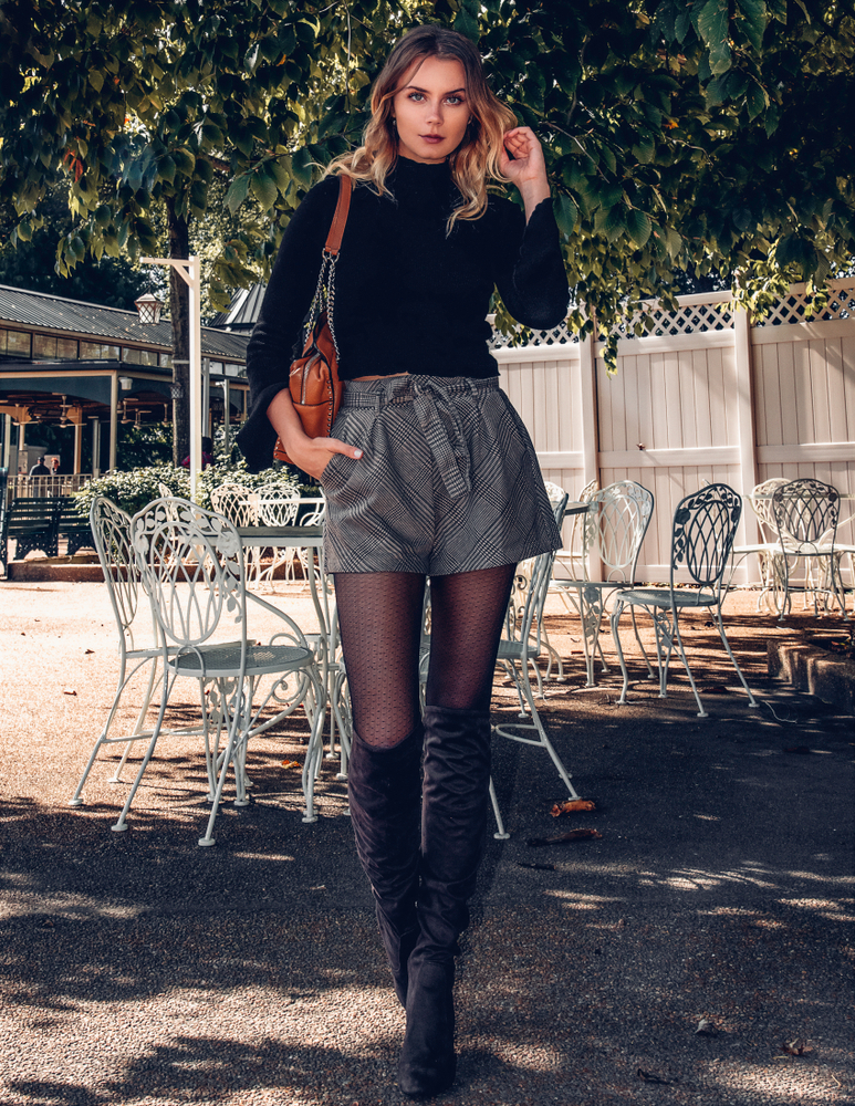 A girl stands on a warm November day outside near patio tables with a turtleneck, shorts, and tights on.