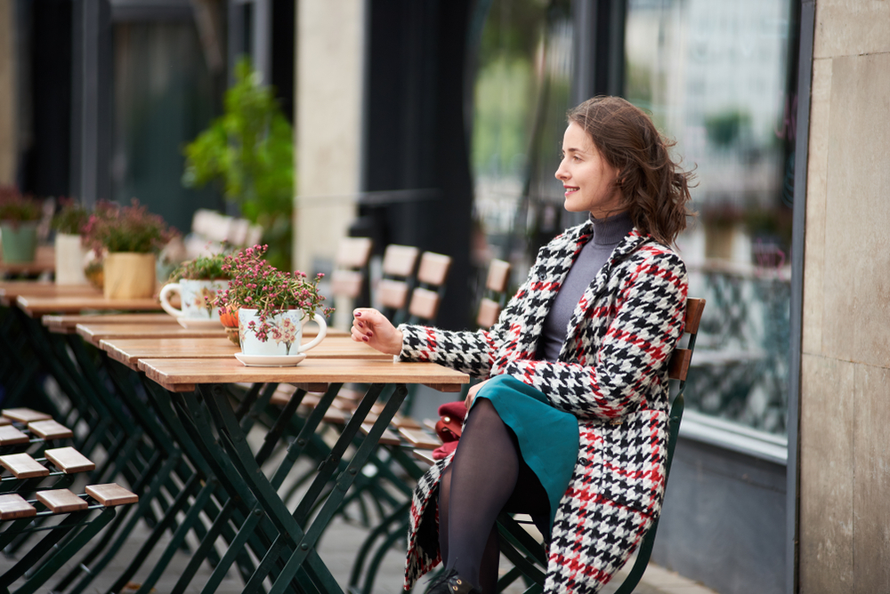 a woman is sitting at a table outside of a cafe in budapest, outfit ideas, she has on a turtle neck, skirt, and a long coat