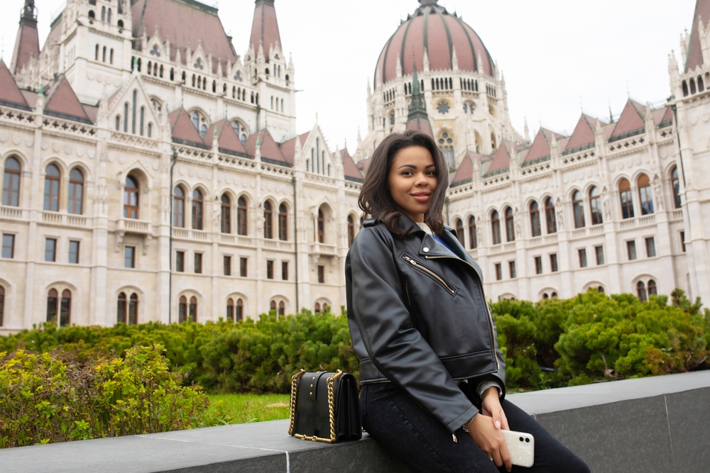 a woman in a leather jacket sits on a stone bench in front of an historic building, her purse is sitting beside her