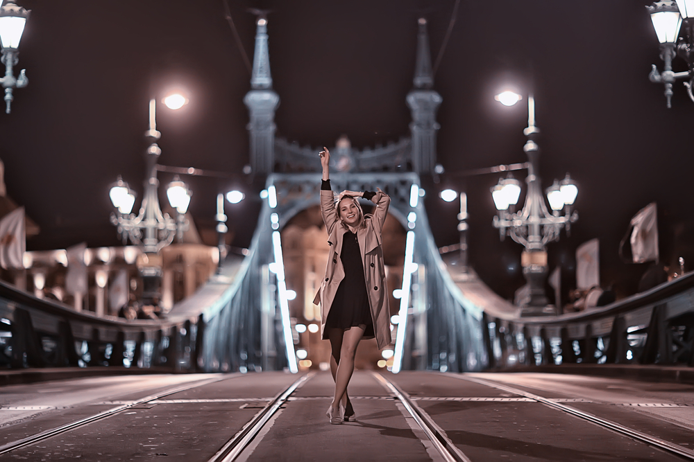 a woman is standing on a bridge at night, she's wearing a black dress and a khaki trench coat over top of it, her arms are raised in the air, what to wear in budapest hungary