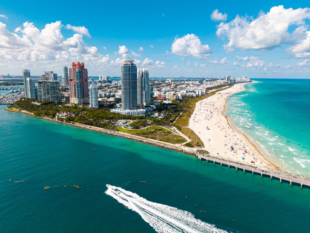 aerial photo of water and beaches with lands in the middle oof the photo with skyscrapers on it, beach chairs and umbrellas are on the beaches, a boat is going through the water in the foreground