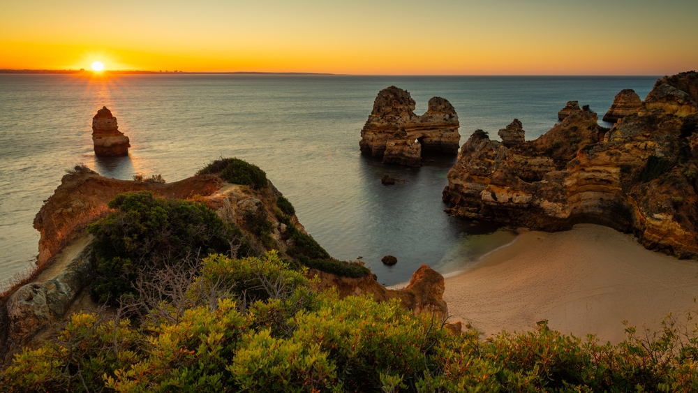 An overhead show of the Algarve in Portugal shows tall, tan rocks, soft sands, and calm waters as the sun sets. 