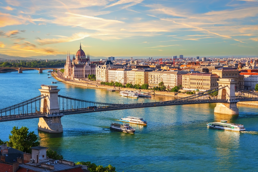 An arial shot of Budapest shows Danbue cruises sailing under tall bridges, the buildings on the coast tall, orange, and vast. 