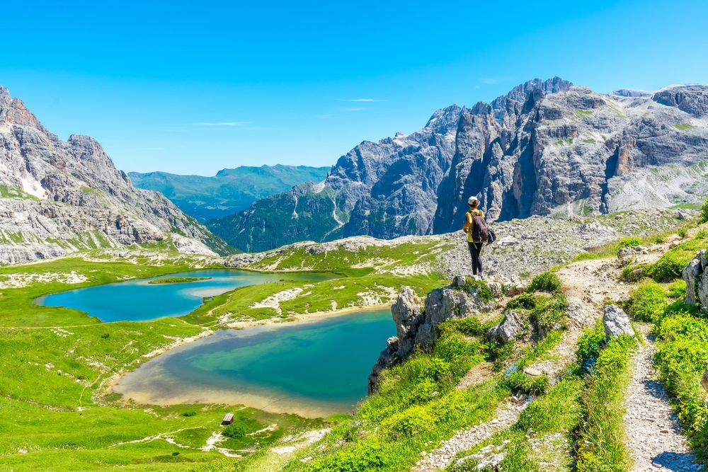 A hiker stands overlooking lakes and greenery in the Dolomites, the view encompassed by rugged peaks, noting it as one of the top places for iconic Europe Trips to Take in 2026. 