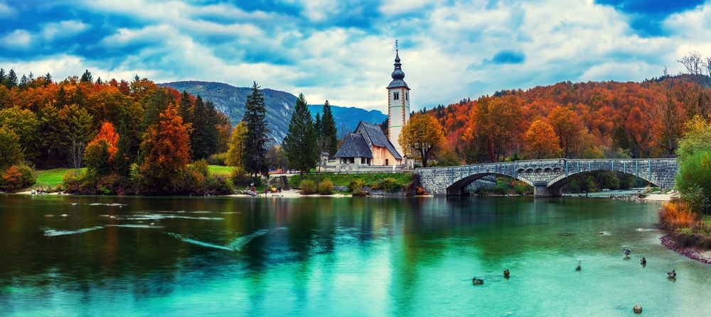 A shot of Slovenia shows green-blue water of a lake, a tall church in the distance, and leaves turning red and orange, welcoming fall. 