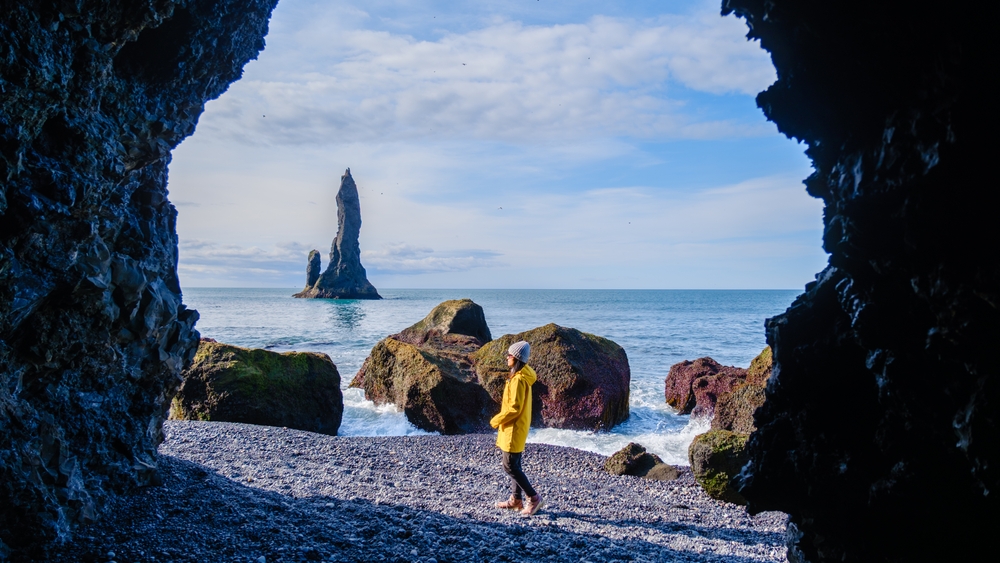 A woman in a yellow coat and beanie walks through black sand in Iceland, framed by the mouth of a cave. 